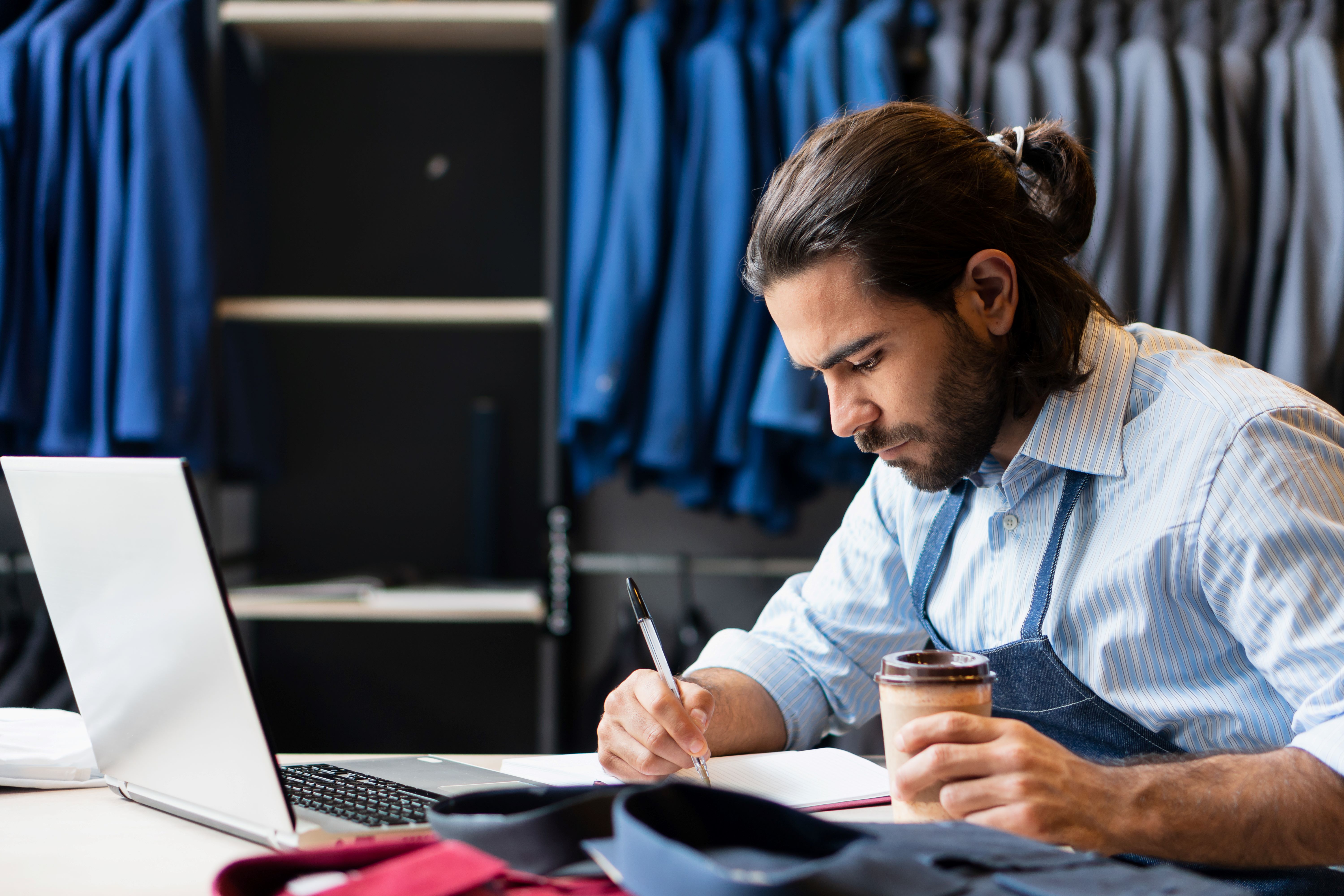 Male tailor writing down notes on paper in front of a laptop, surrounded by suits