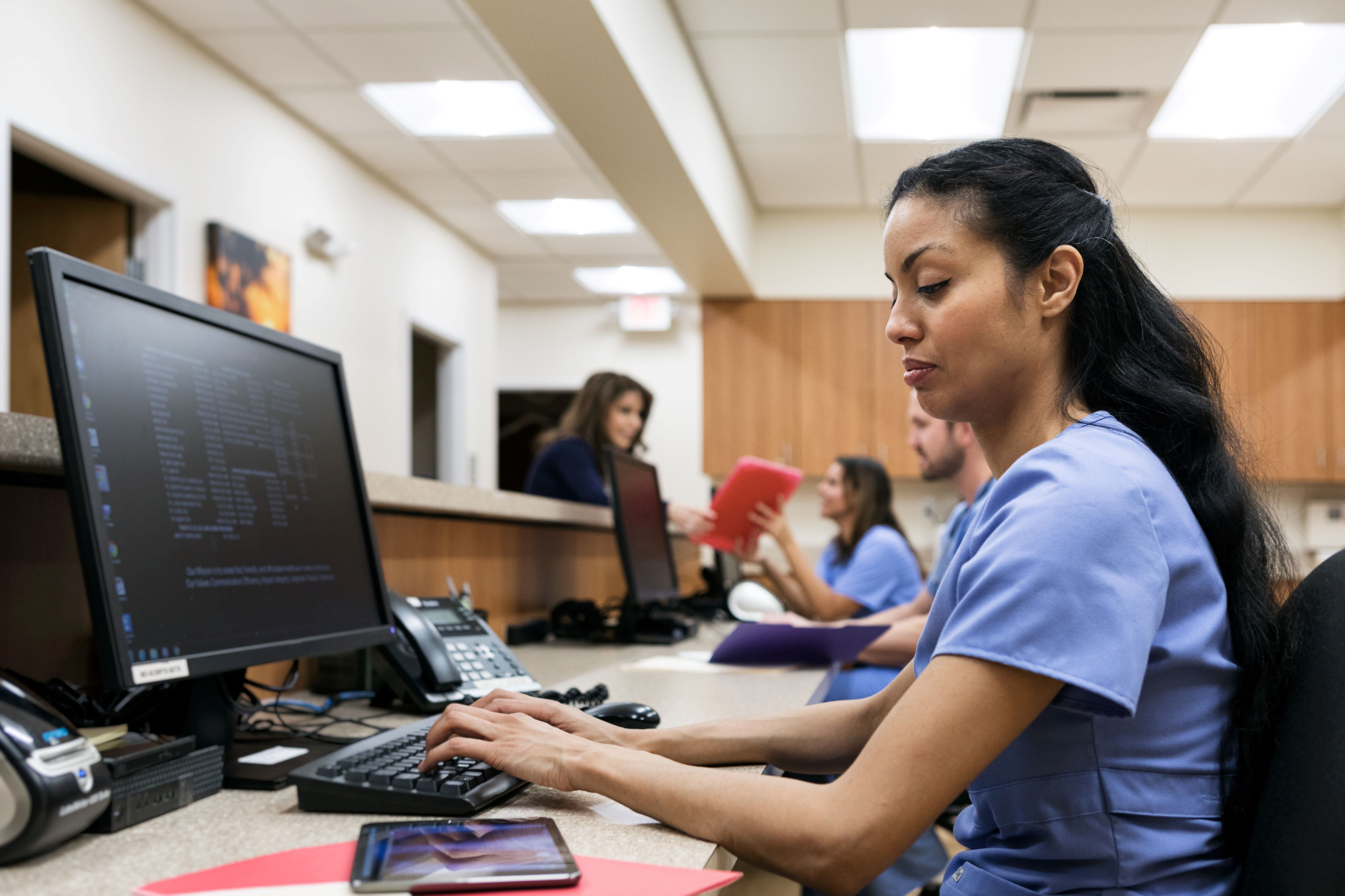 Medical professional sat at desk entering data from tablet into computer