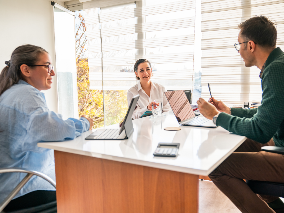 Three people in a bright office sitting around a table with laptops and a calculator, engaged in a discussion.