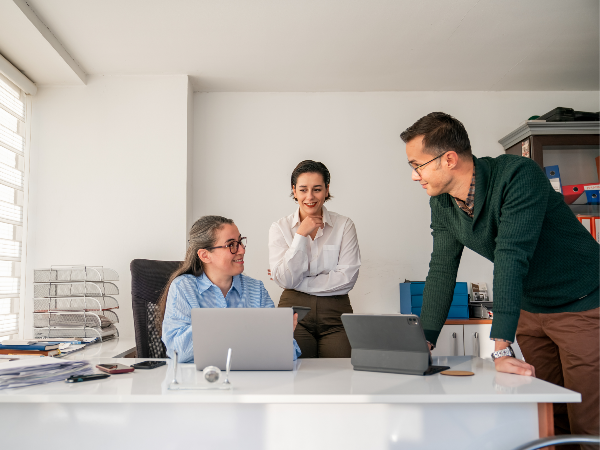 Three people in a modern office setting collaborating around a desk with laptops and documents.