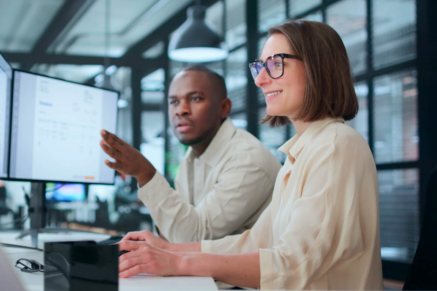 Smiling accountant wearing glasses sitting next to coworker in modern office looking at multiple screens showing invoices