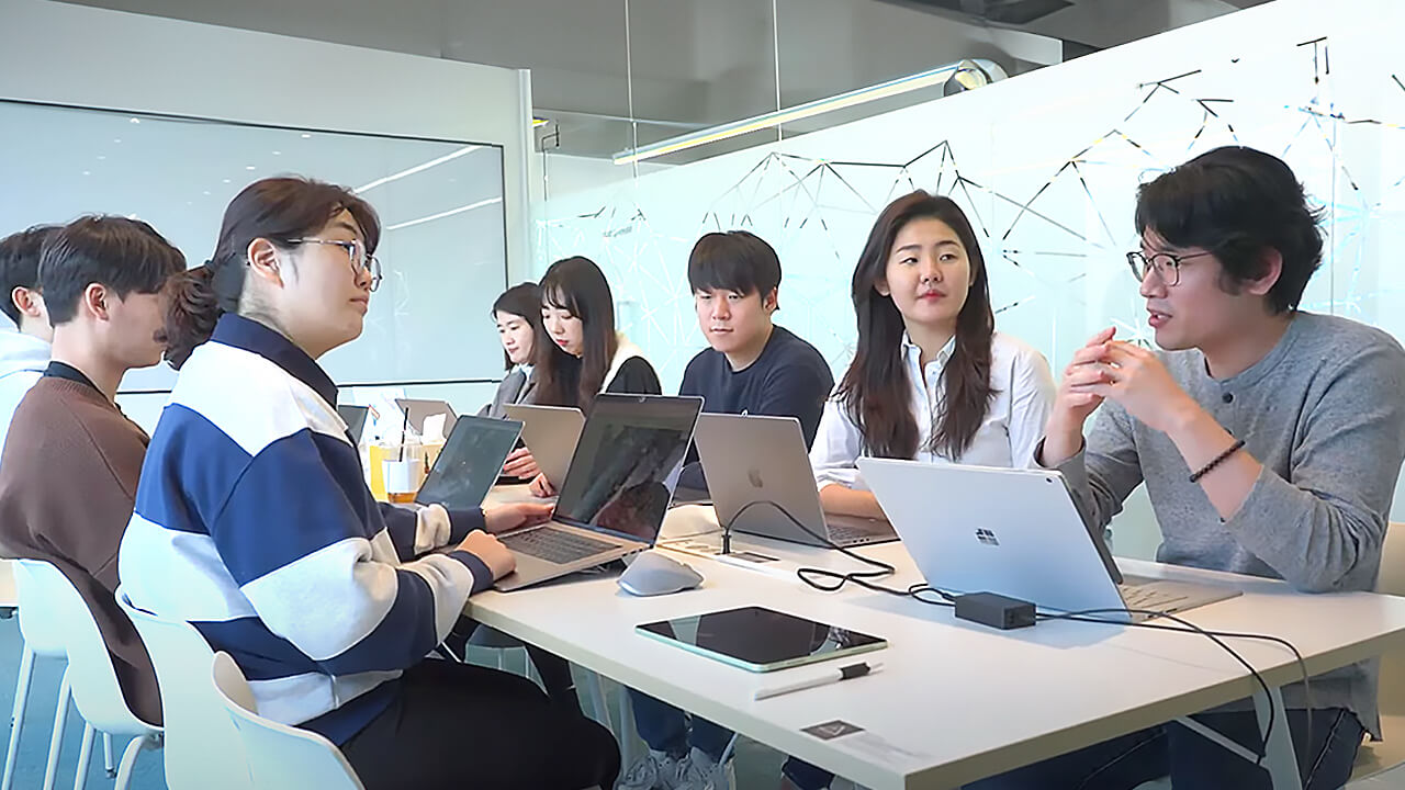 A group of people collaborating during a team meeting in a modern office environment, seated around a table with laptops and digital devices.