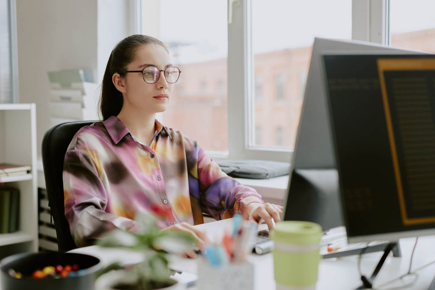 Technical expert wearing colorful shirt while looking at a computer screen in office building overlooking buildings