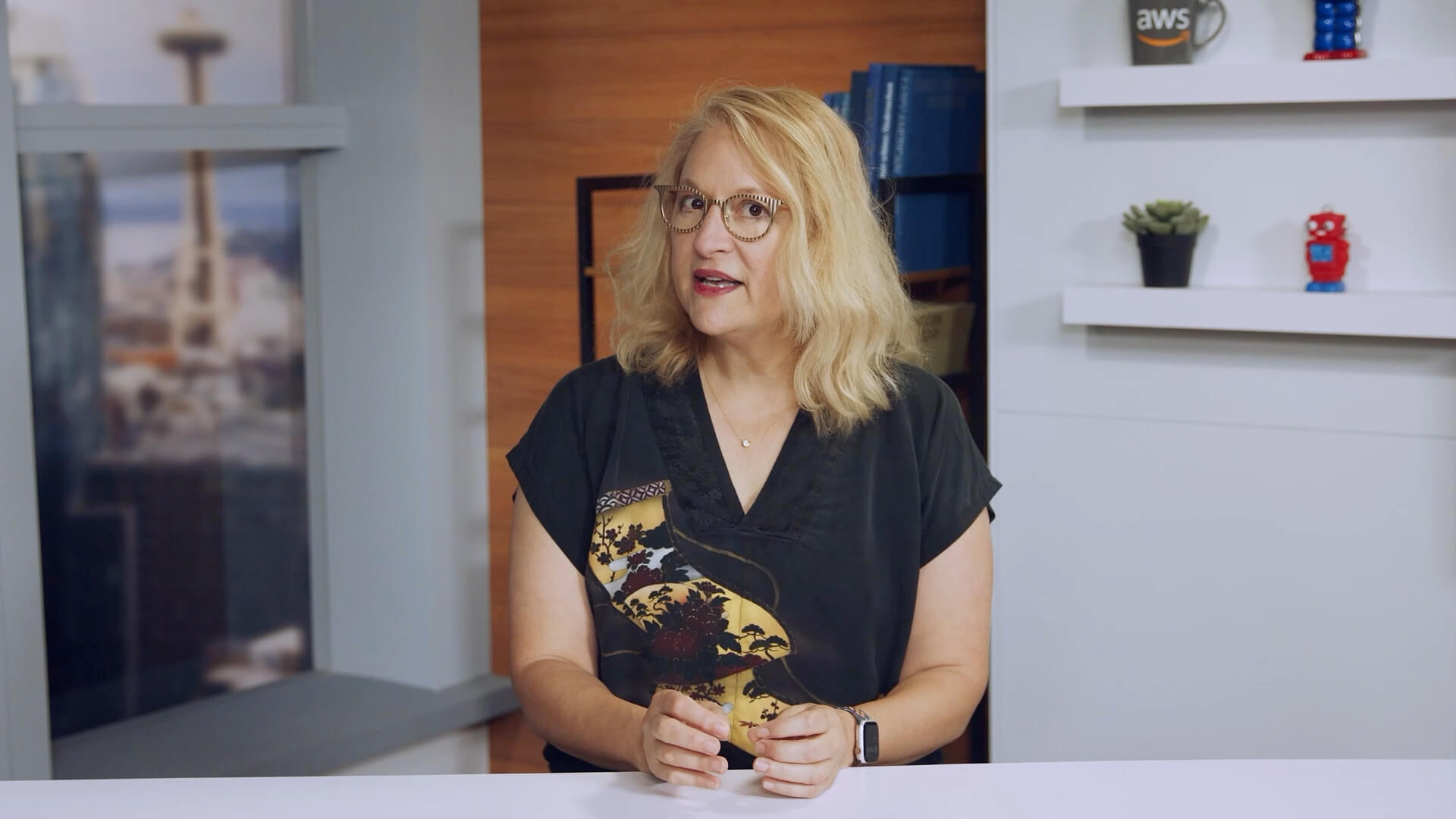 A woman is presenting information about data collection and analysis in a modern AWS office environment. The setting features shelves with AWS-branded items and a window with a view of the city, indicating a professional and technology-focused atmosphere.