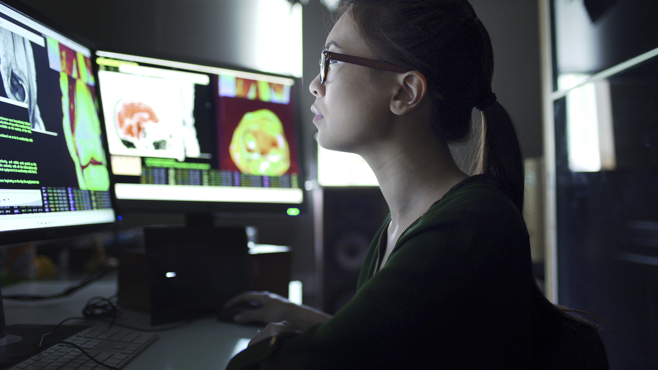 Close up image of a young asian woman working at a desk with multiple monitors.