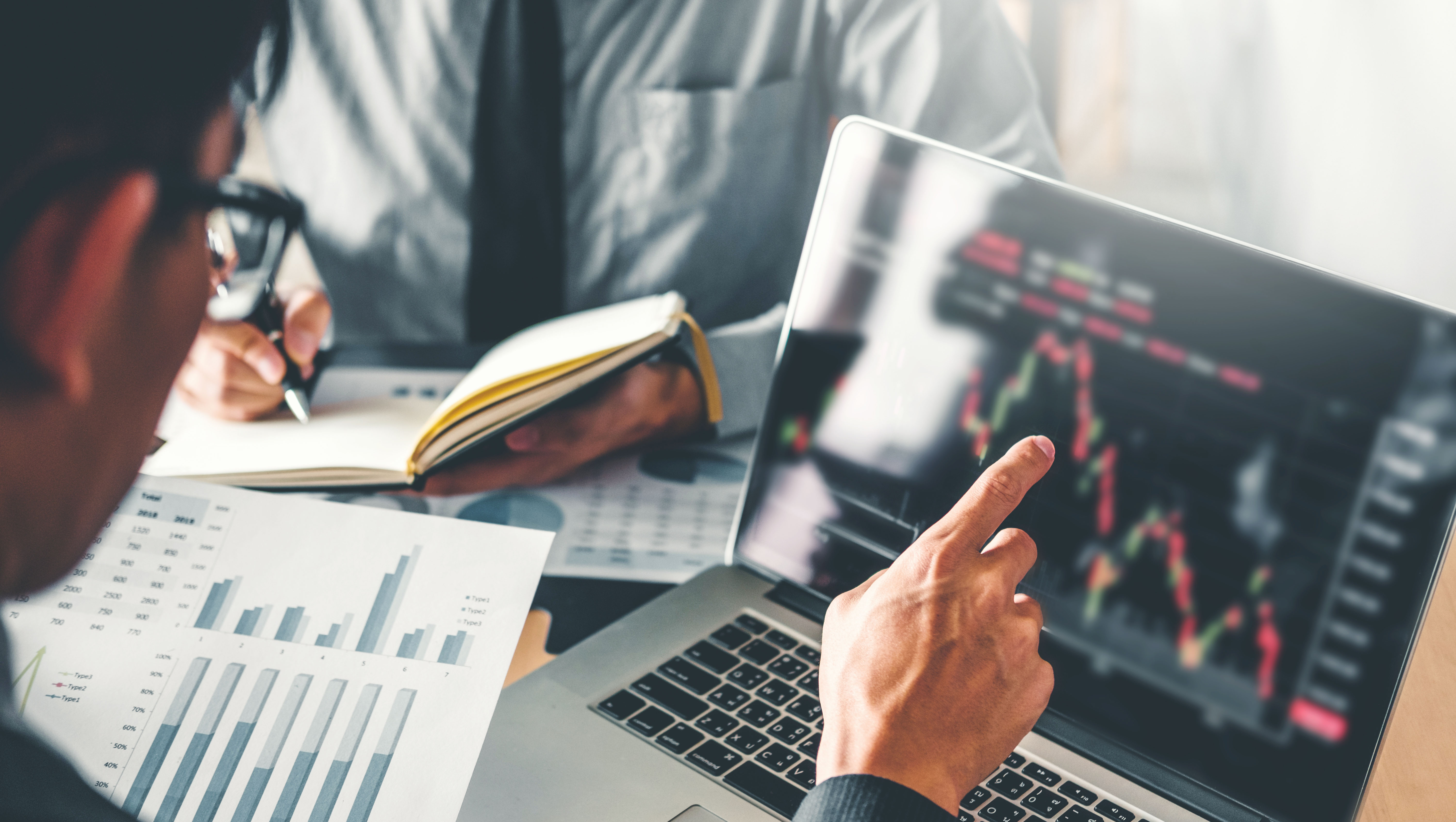 Two professionals review financial data and analytics, with bar charts on paper and stock market graphs displayed on a laptop screen.