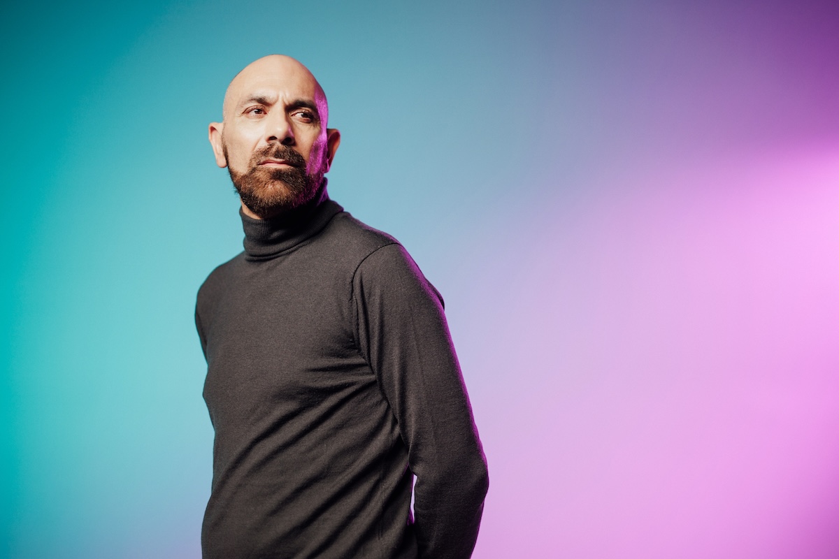 A person wearing a dark turtleneck sweater against a gradient background of teal and pink.