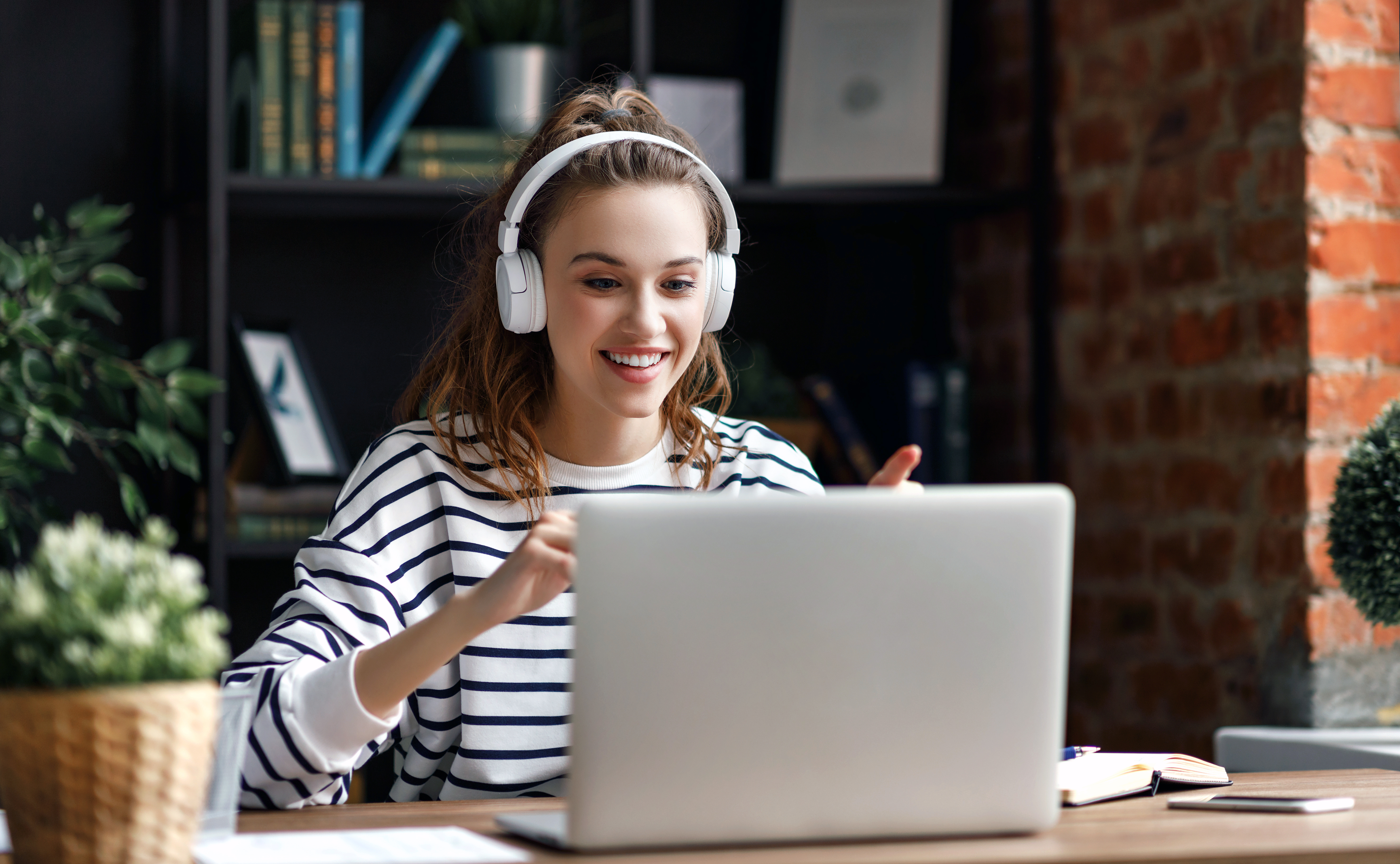 Happy millennial female in casual clothes and wireless headphones gesticulating while sharing news with friend having video call via laptop at table in loft office