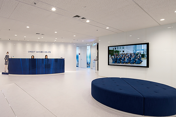 Reception area of Credit Saison office featuring a blue reception desk, a large modern circular seating area, and a wall-mounted screen displaying a group photo.