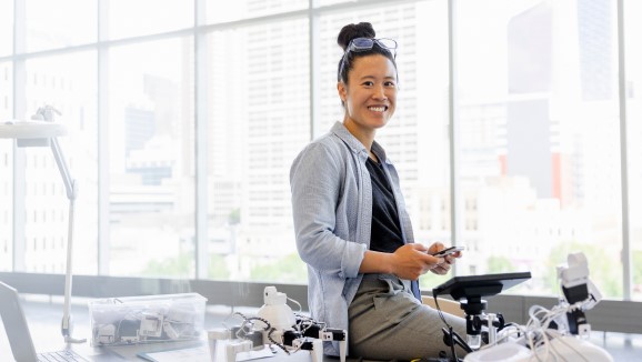A smiling engineer sitting at a desk surrounded by robotics equipment and electronic devices in a modern office environment with large windows and a cityscape background.