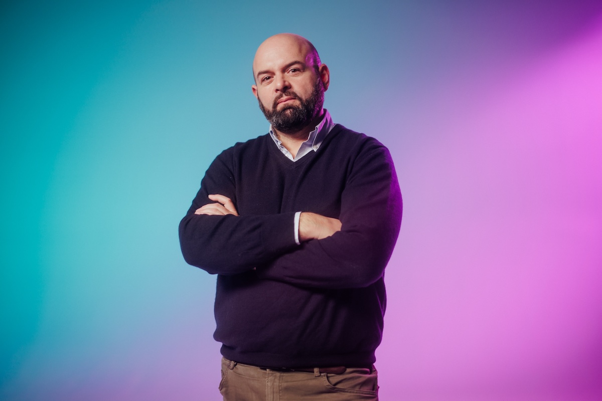 A bald man with a beard stands with his arms crossed, wearing a dark sweater and light pants, in front of a vibrant background with blue and pink gradient lighting.
