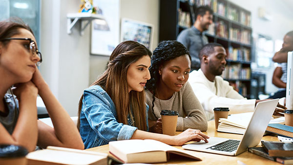 A group of students collaborating and working together in a library setting, using a laptop and surrounded by books and coffee cups.