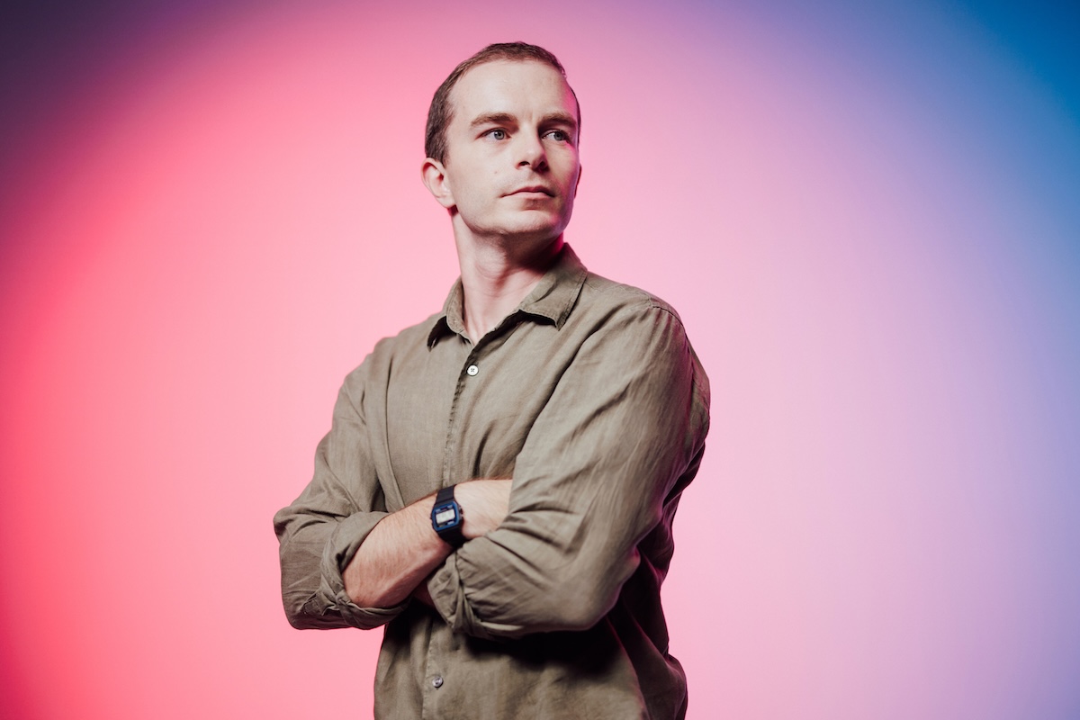 A person standing with arms crossed, looking to the side, with a colorful pink and blue gradient background.