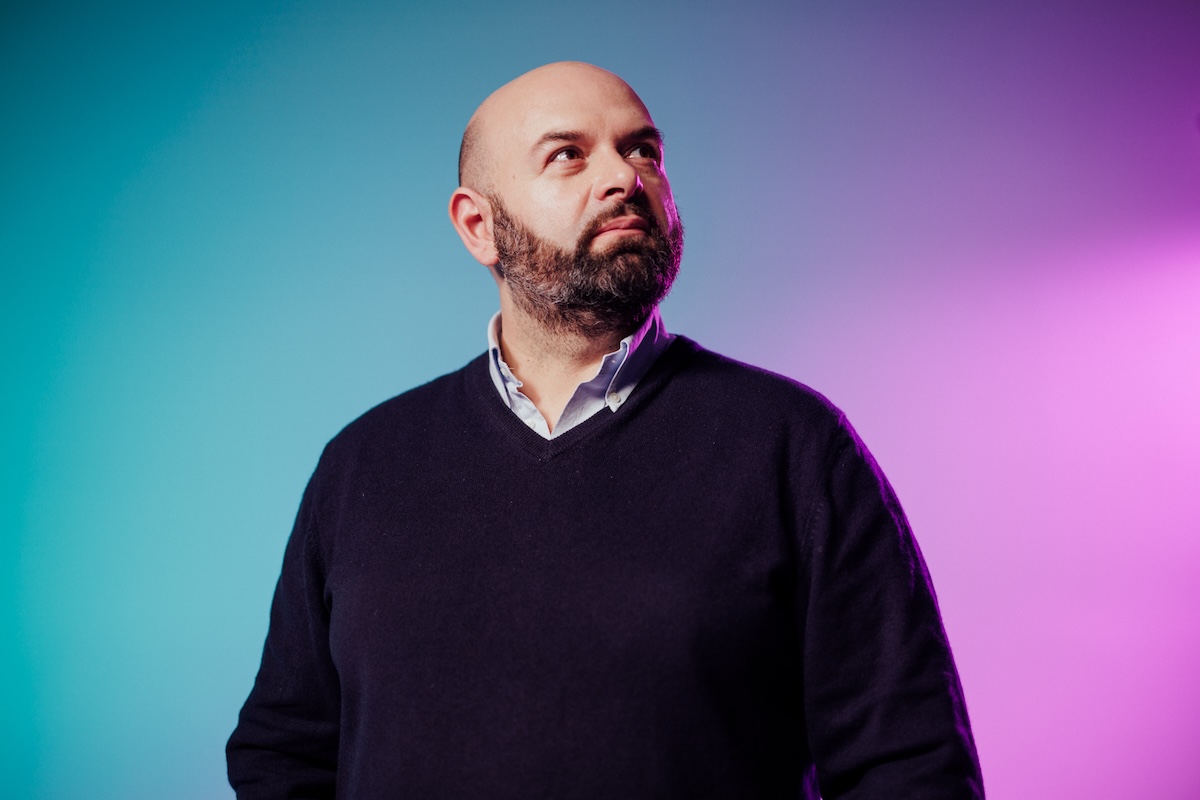 A portrait of a bearded man in a dark sweater and collared shirt, standing against a colorful gradient background with blue and purple lighting.