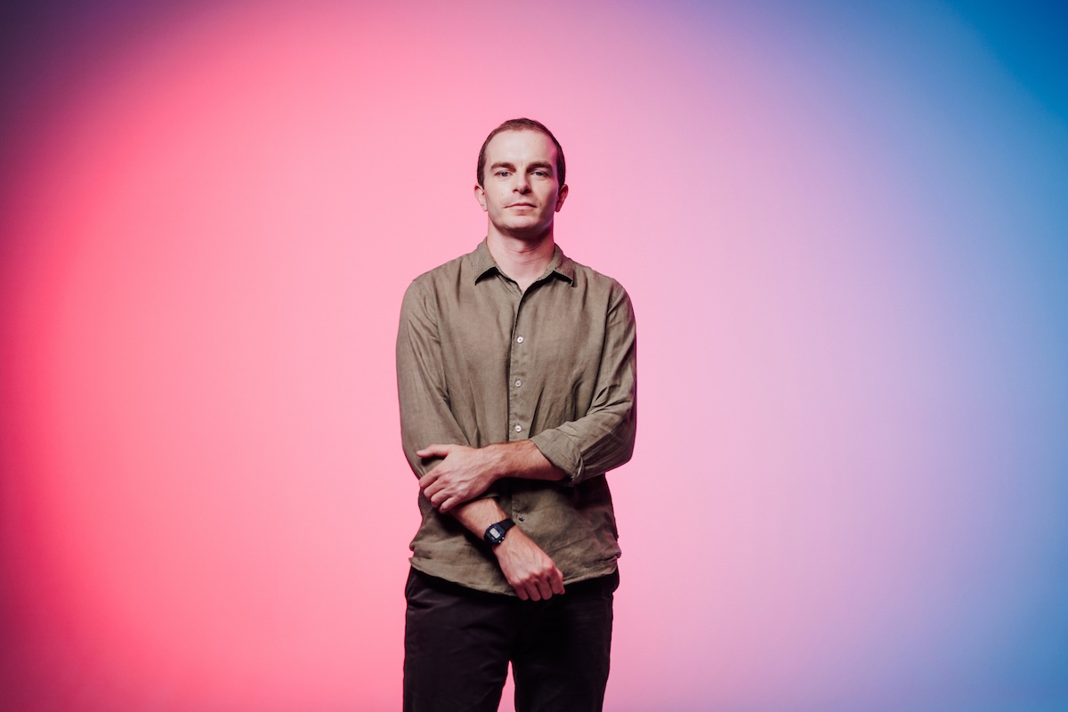 A man stands with arms crossed in front of a vibrant gradient background transitioning from pink to blue.