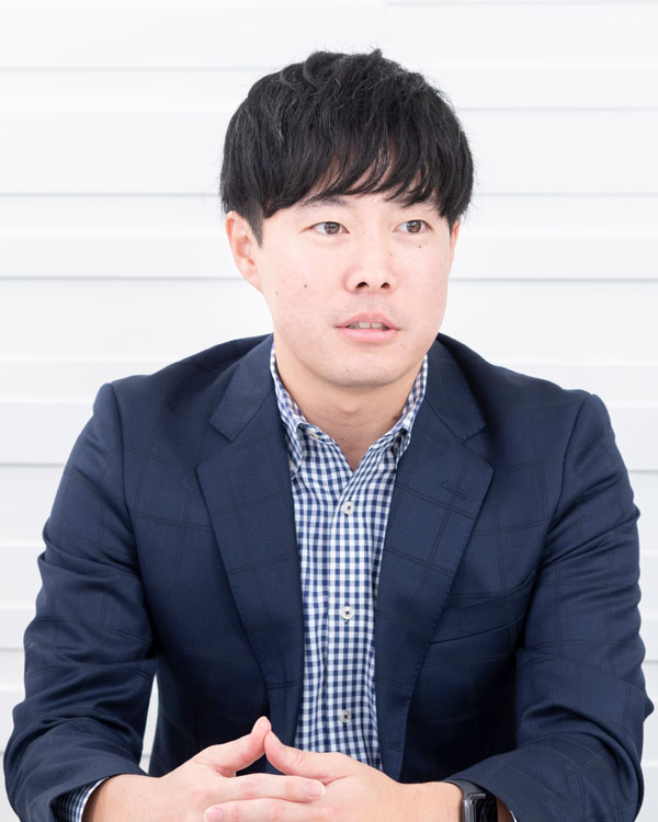 A person wearing a navy blazer over a checkered shirt, seated with hands clasped, against a white background.
