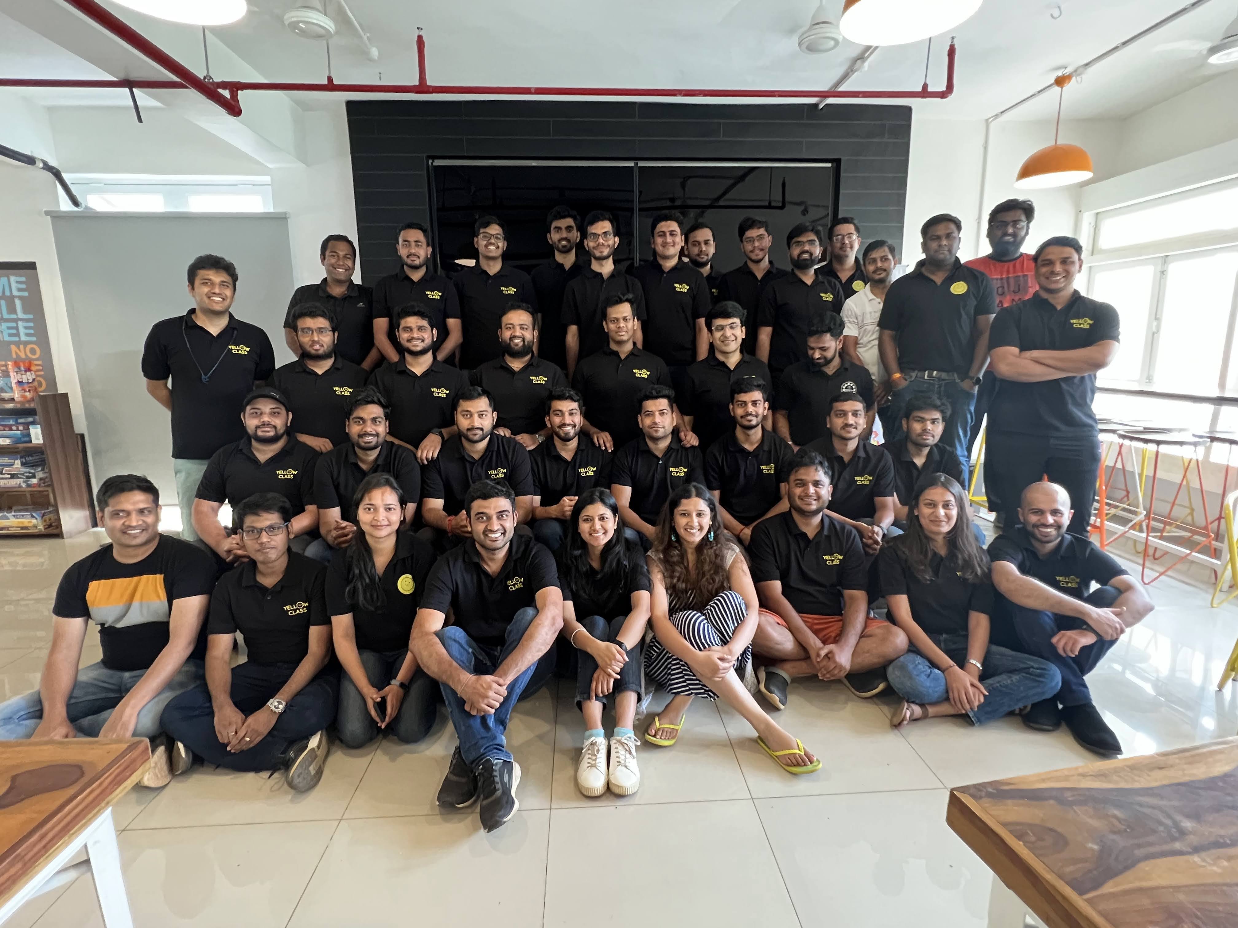 A large group of people, all wearing matching black 'Yellow Class' shirts, pose together for a team photo in a modern office setting with bright natural light.