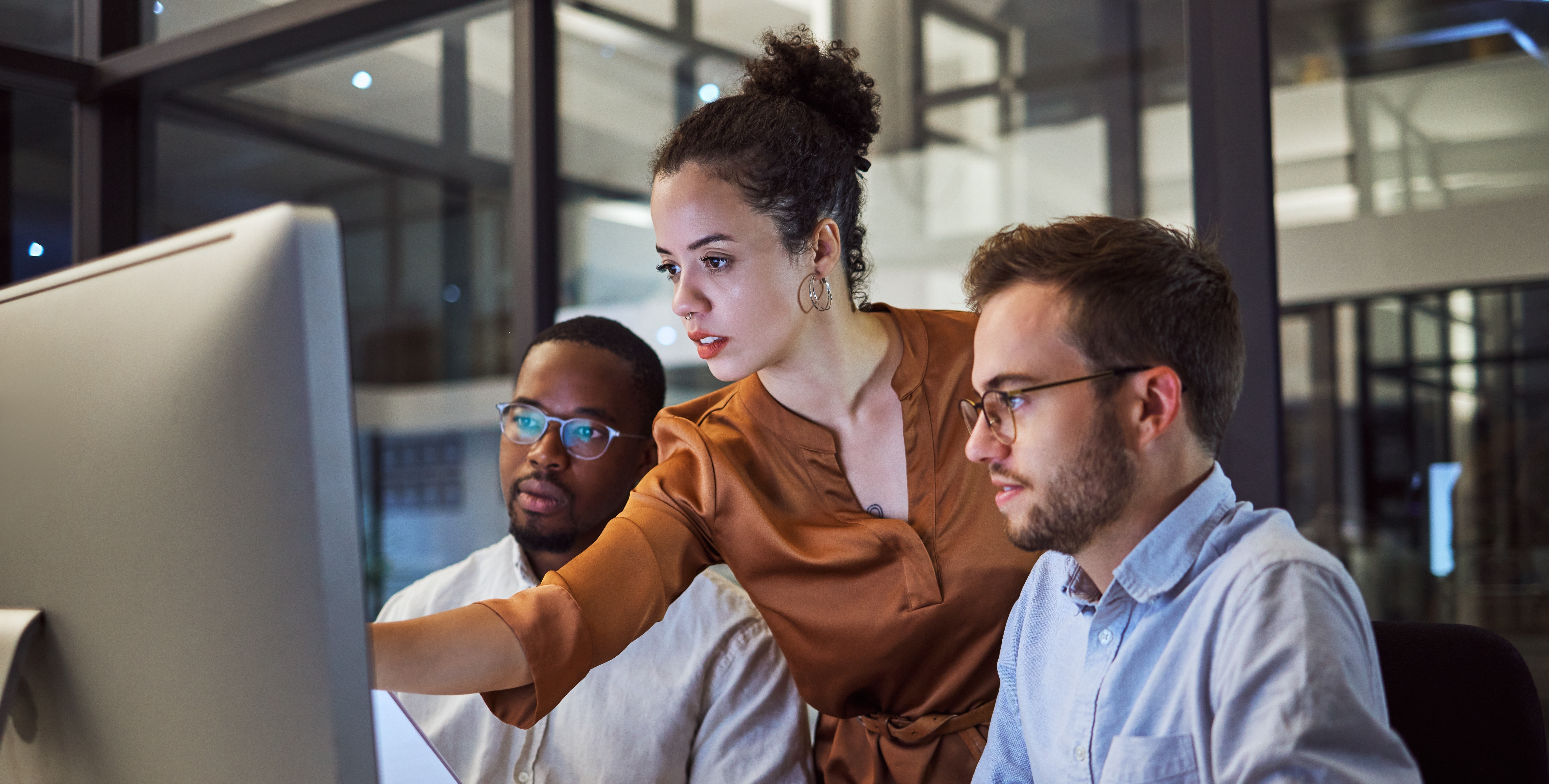 A diverse group of three colleagues working together at a computer workstation in a modern office environment, analyzing data or discussing a project.