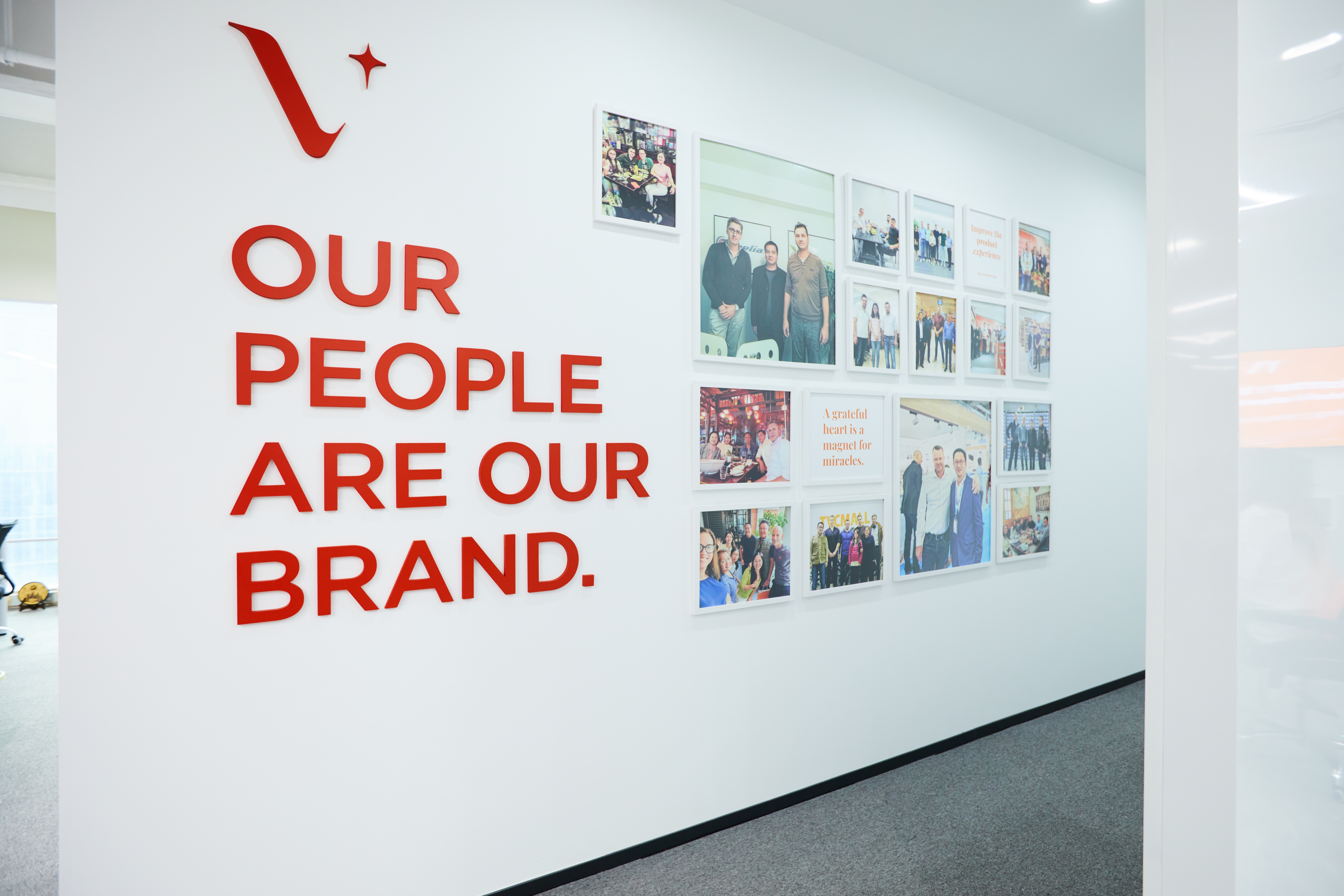 A white office wall with red text reading "Our People Are Our Brand" and a collage of framed photos showcasing team moments and quotes.