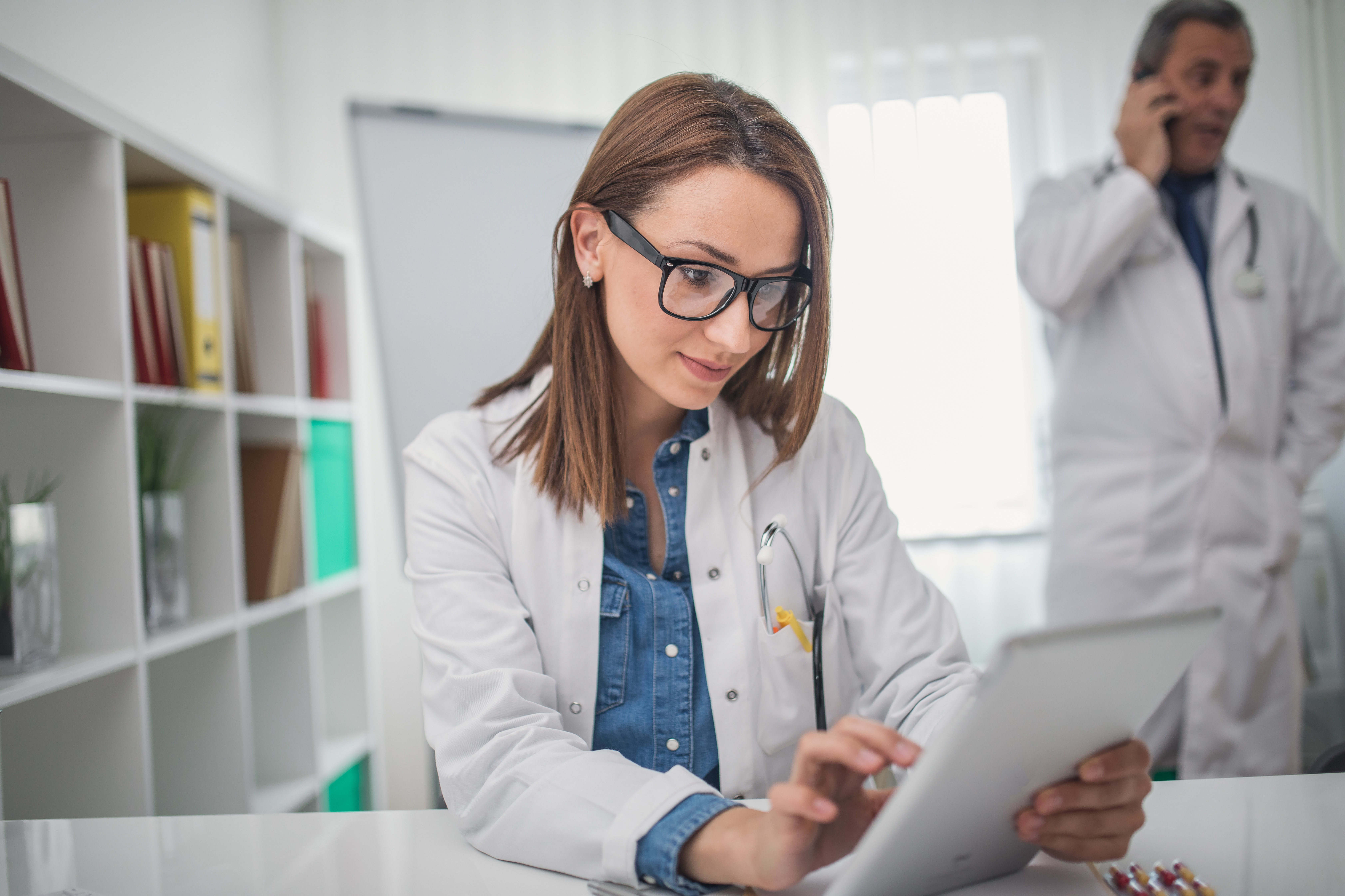 A medical professional in a white coat using a tablet at a desk, with shelves and another doctor in the background.