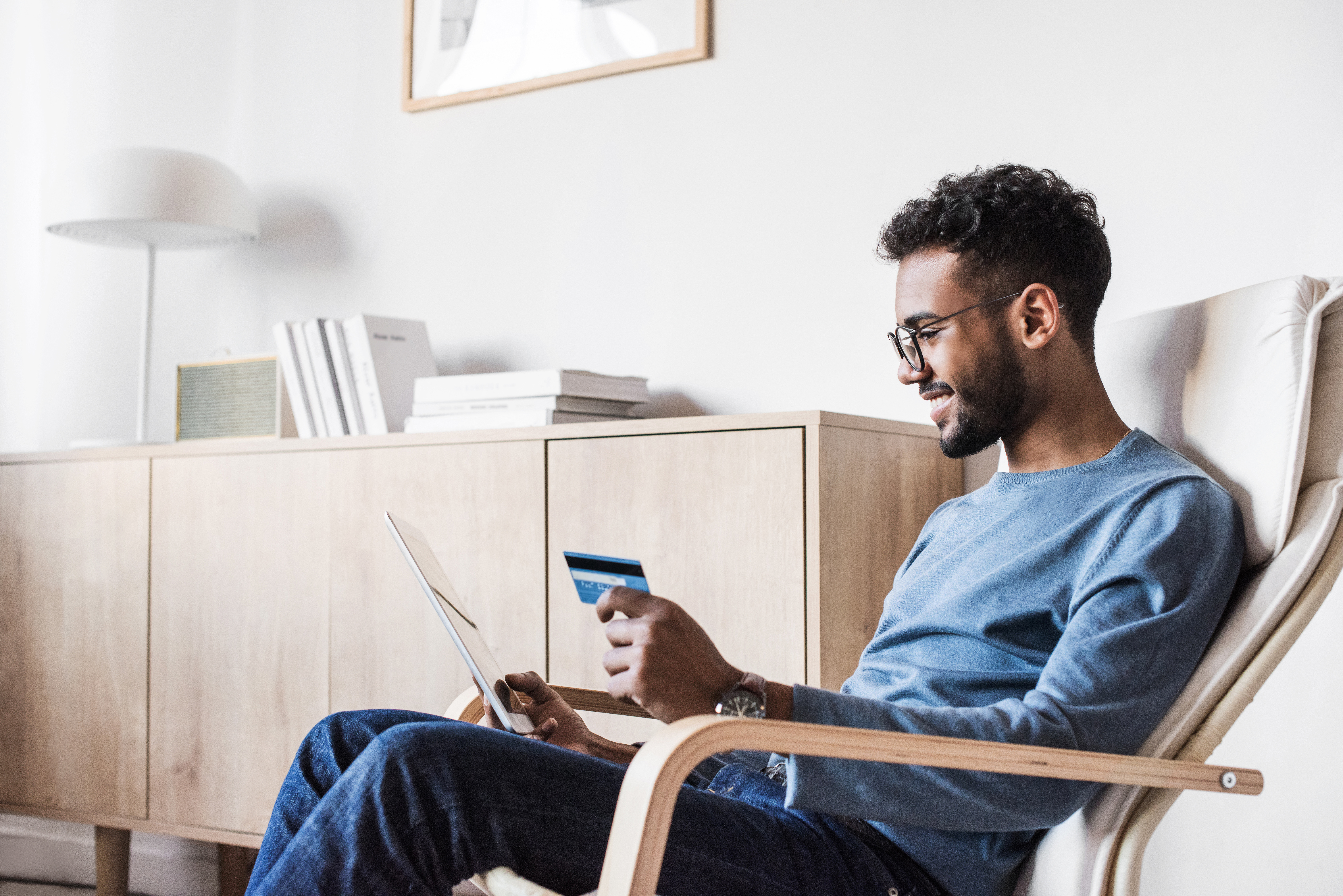 A man sitting comfortably in a modern living room, smiling while making an online purchase using a tablet and holding a credit card.