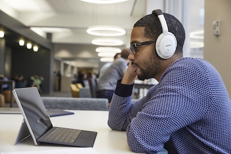 A person wearing headphones sits at a table in a modern office environment, looking at a tablet or laptop device. Other people and circular ceiling lights are visible in the background.