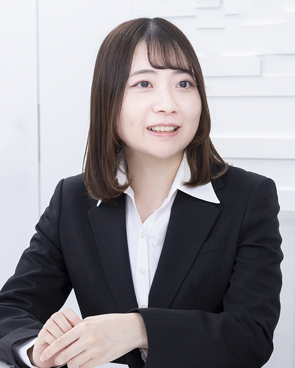A person wearing a black suit and white collared shirt, seated in a bright, modern setting.