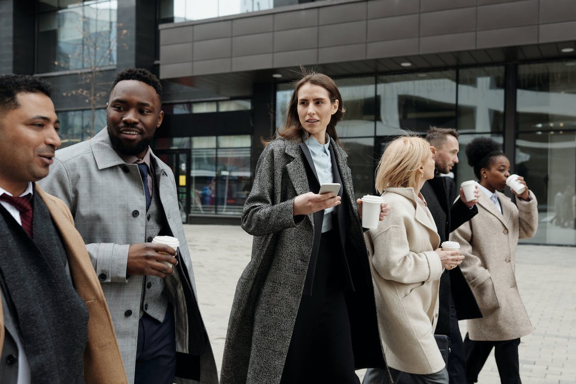 Group of people in formal coats walking outdoors, holding coffee cups and a smartphone, with a modern building in the background.