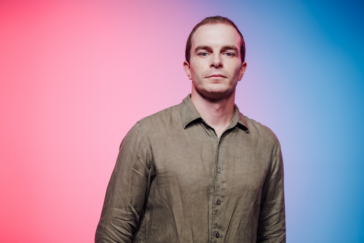 A headshot of a person wearing a button-up shirt, posed in front of a gradient background transitioning from pink on the left to blue on the right.