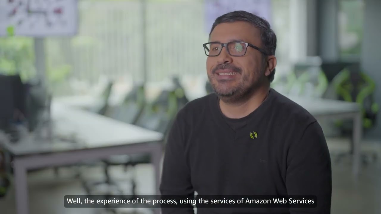 YouTube video thumbnail showing an interview with a person discussing their experience using Amazon Web Services, with office background and English subtitle: 'Well, the experience of the process, using the services of Amazon Web Services.'