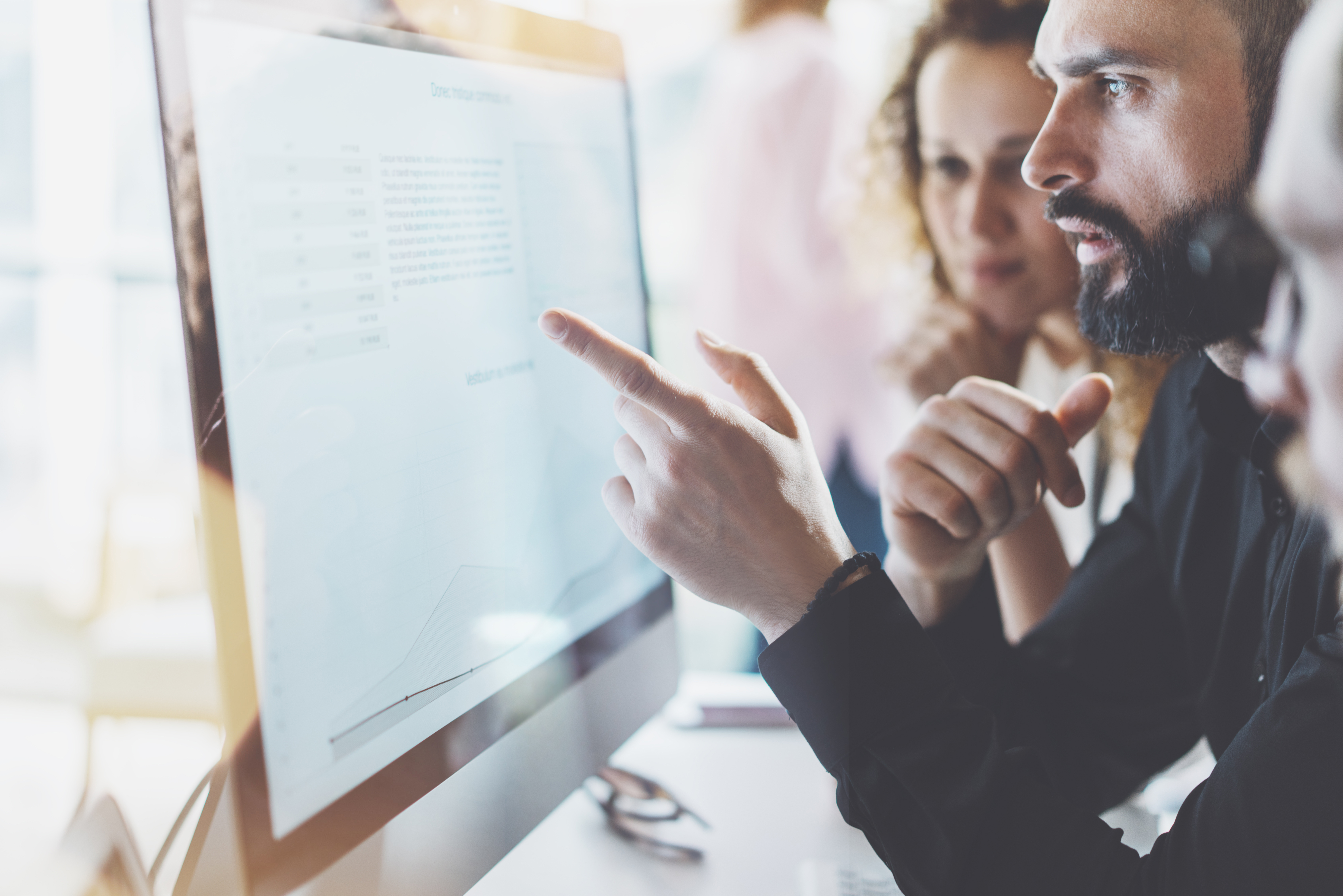 A group of people closely analyzing data on a large computer monitor, collaborating and discussing information displayed on the screen in a modern office setting.