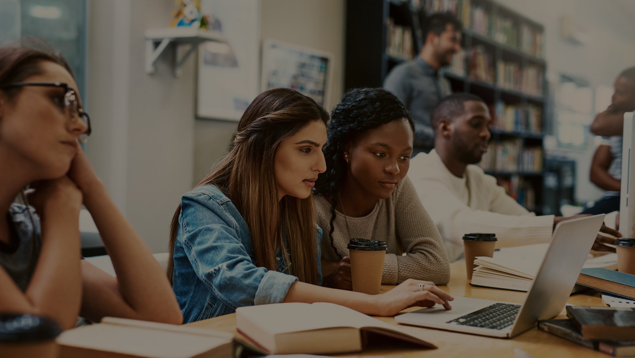 A group of students working together at a table in a library, using laptops and surrounded by open books and coffee cups. The environment appears collaborative and focused on study or group work.