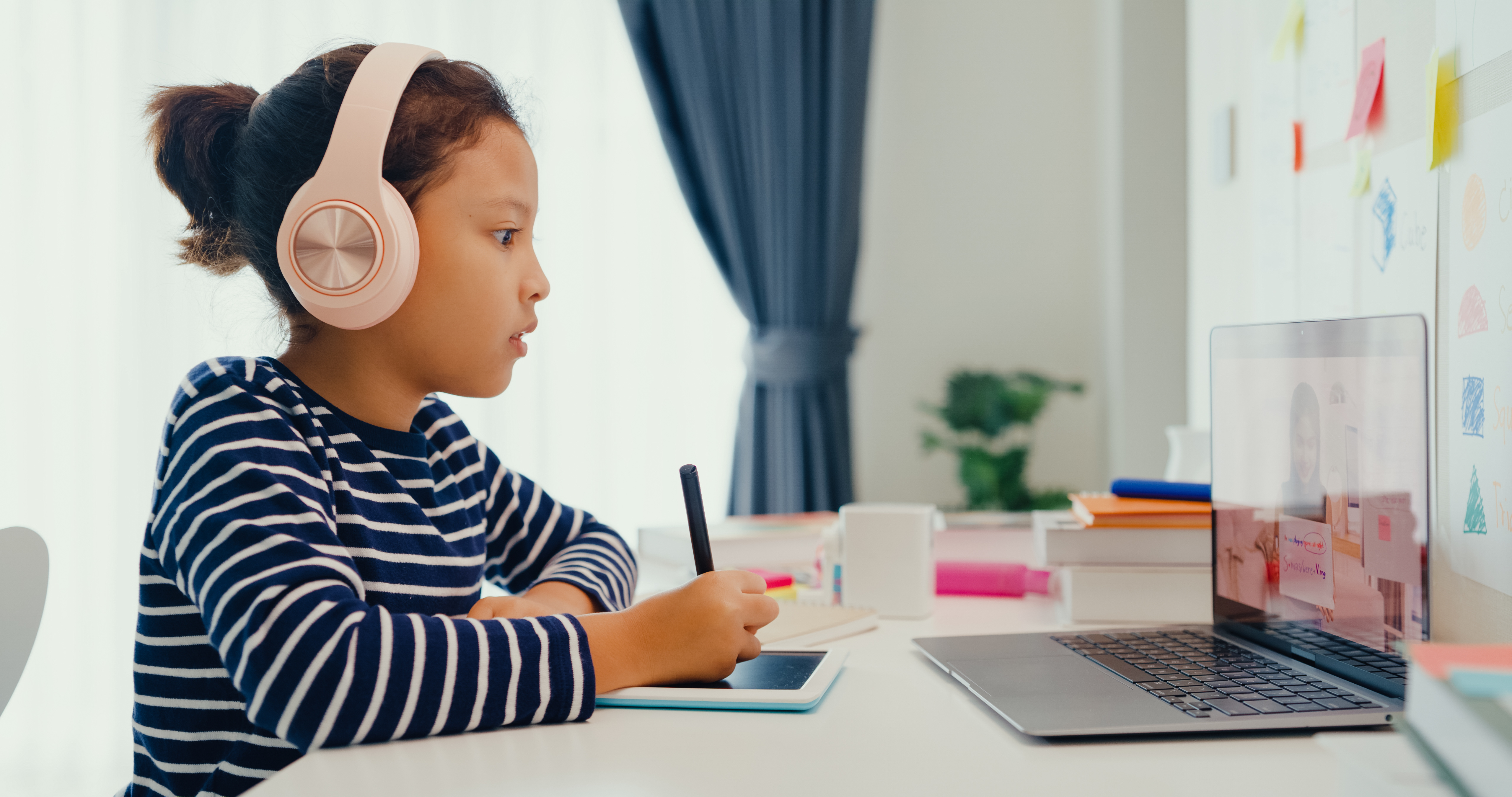 A child wearing headphones sits at a desk, attentively watching an educational video on a laptop while taking notes with a stylus, representing remote learning or online education.