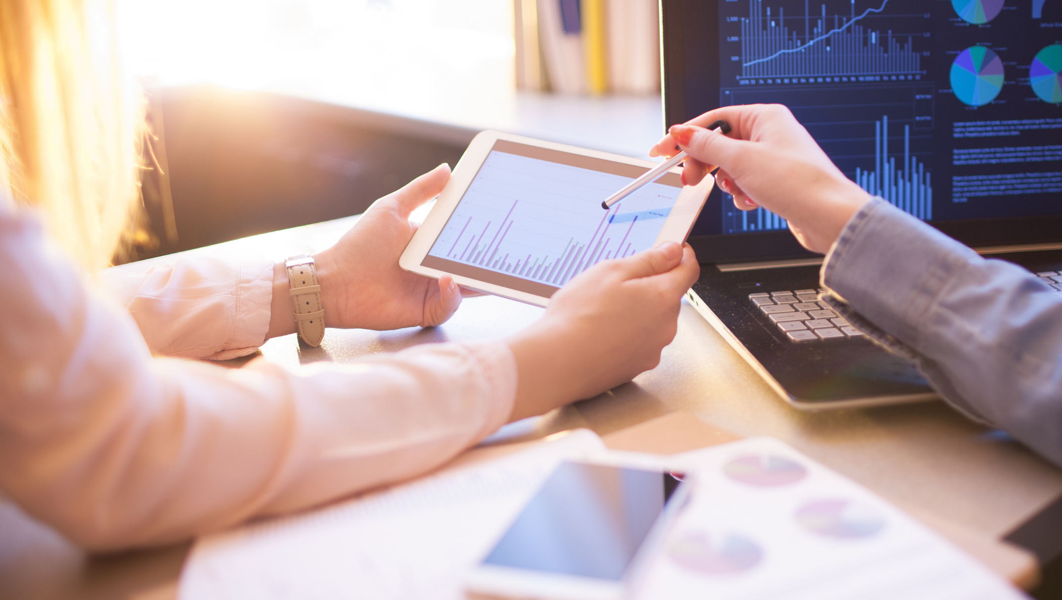 Two people reviewing digital charts and data analytics on a tablet and laptop in a sunlit office environment, illustrating teamwork and business analysis.