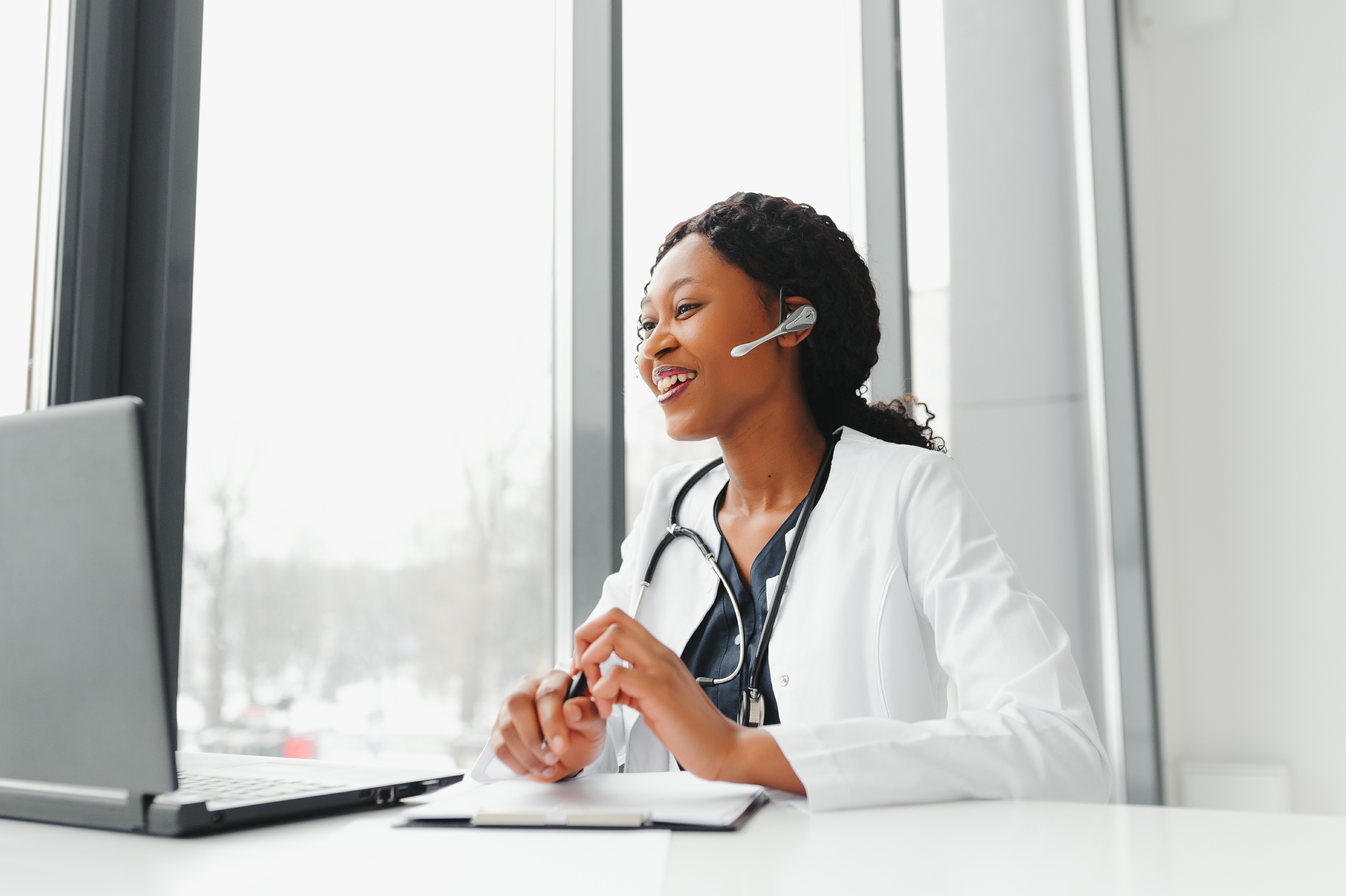 A doctor wearing a headset and white coat with a stethoscope, sitting at a desk with a laptop, participates in a telemedicine video call in a bright office environment.