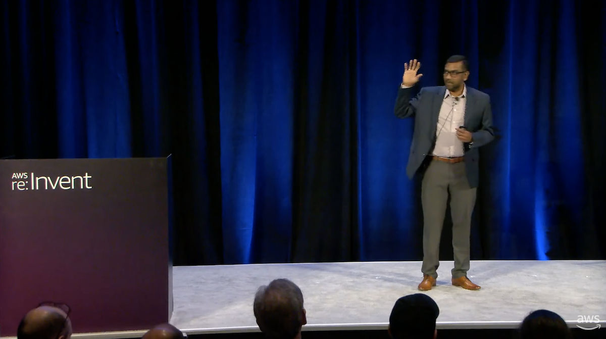 A speaker on stage at AWS re:Invent, gesturing with one hand raised, standing in front of a blue curtain.