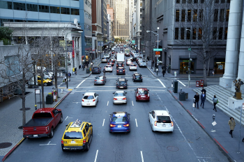 A view of a busy downtown city street with multiple cars and taxis in traffic, surrounded by tall office buildings and pedestrians on the sidewalks.