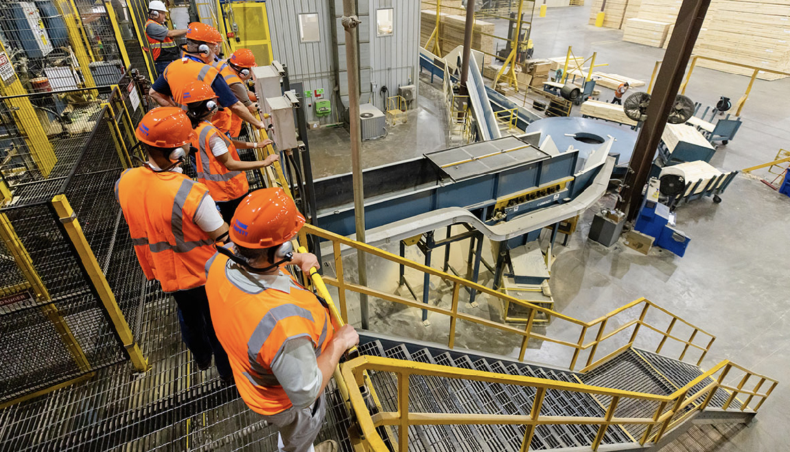 A group of workers wearing orange safety vests and hardhats touring a Georgia-Pacific factory, observing industrial machinery from a stairway inside a manufacturing facility.
