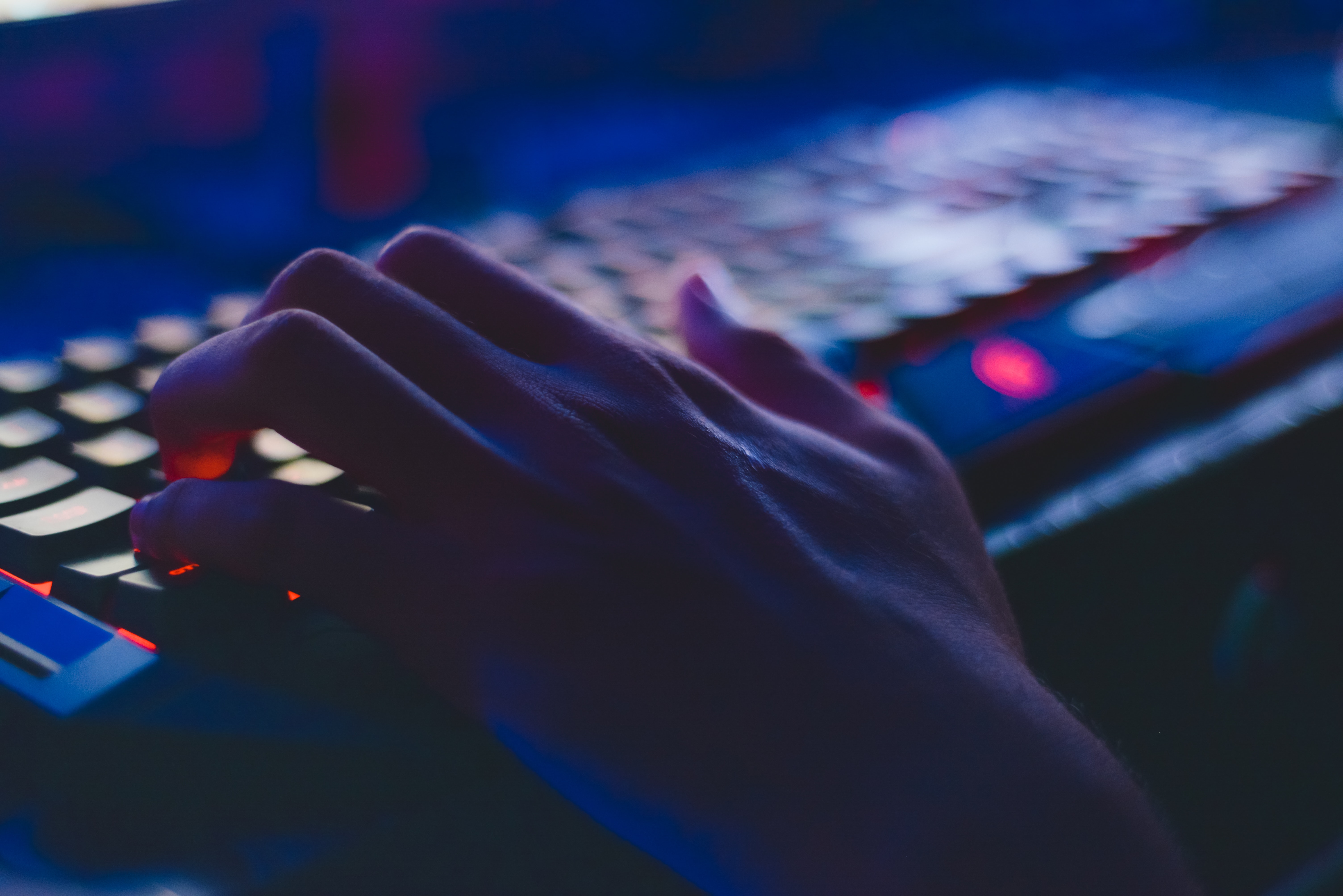 A closeup of a hand resting on a colorful RGB-backlit gaming keyboard in a dark, blue-lit environment.