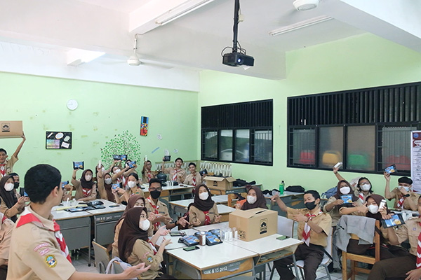 A group of students in uniform attending an offline classroom training session. They are seated at tables, some holding up materials, and most are wearing masks. The classroom is well-lit with educational decorations on the walls, and a teacher is engaging with the students at the front. This scene is part of Rumah Siap Kerja's offline training initiatives.