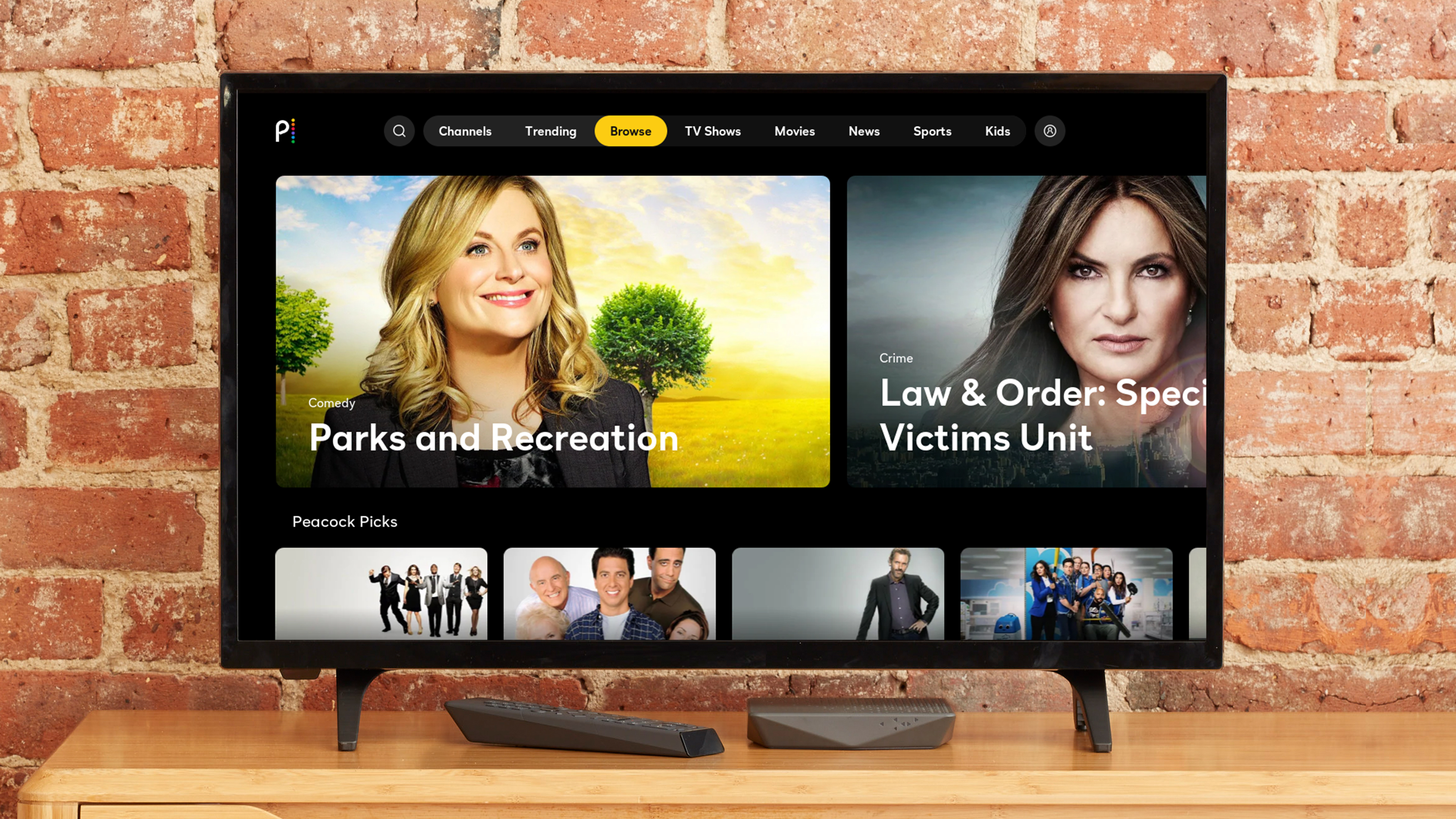 A television screen showing the Peacock streaming service interface, featuring 'Parks and Recreation' and 'Law & Order: Special Victims Unit' as highlighted shows. The TV is placed on a wooden stand against a brick wall.