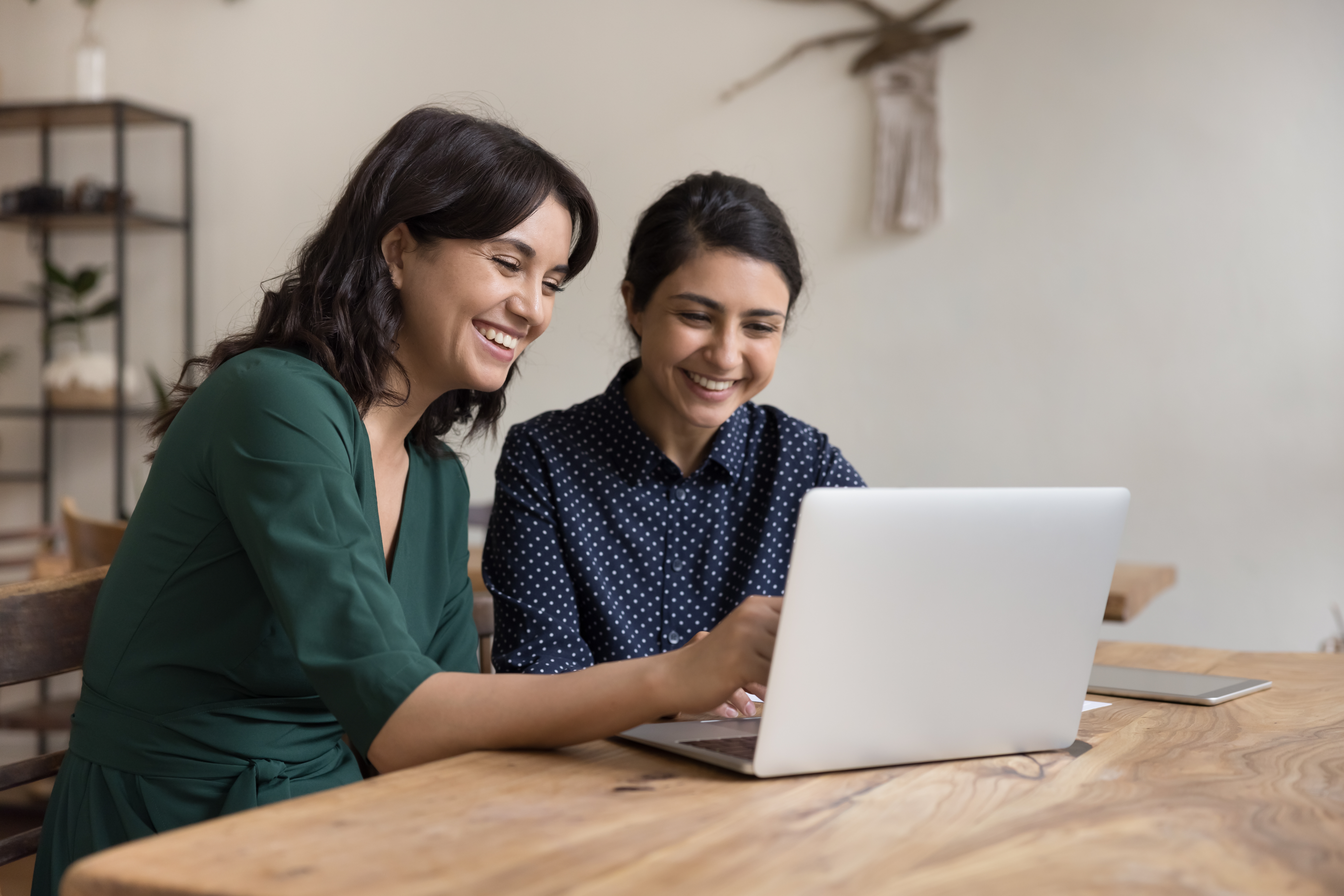 Two cheerful diverse office workers women cooperating on project, sitting at work desk with laptop, looking at screen, smiling, laughing. Female mentor teaching Indian student, pointing at monitor; Shutterstock ID 2238906761; purchase_order: phidias case study; job: AWS