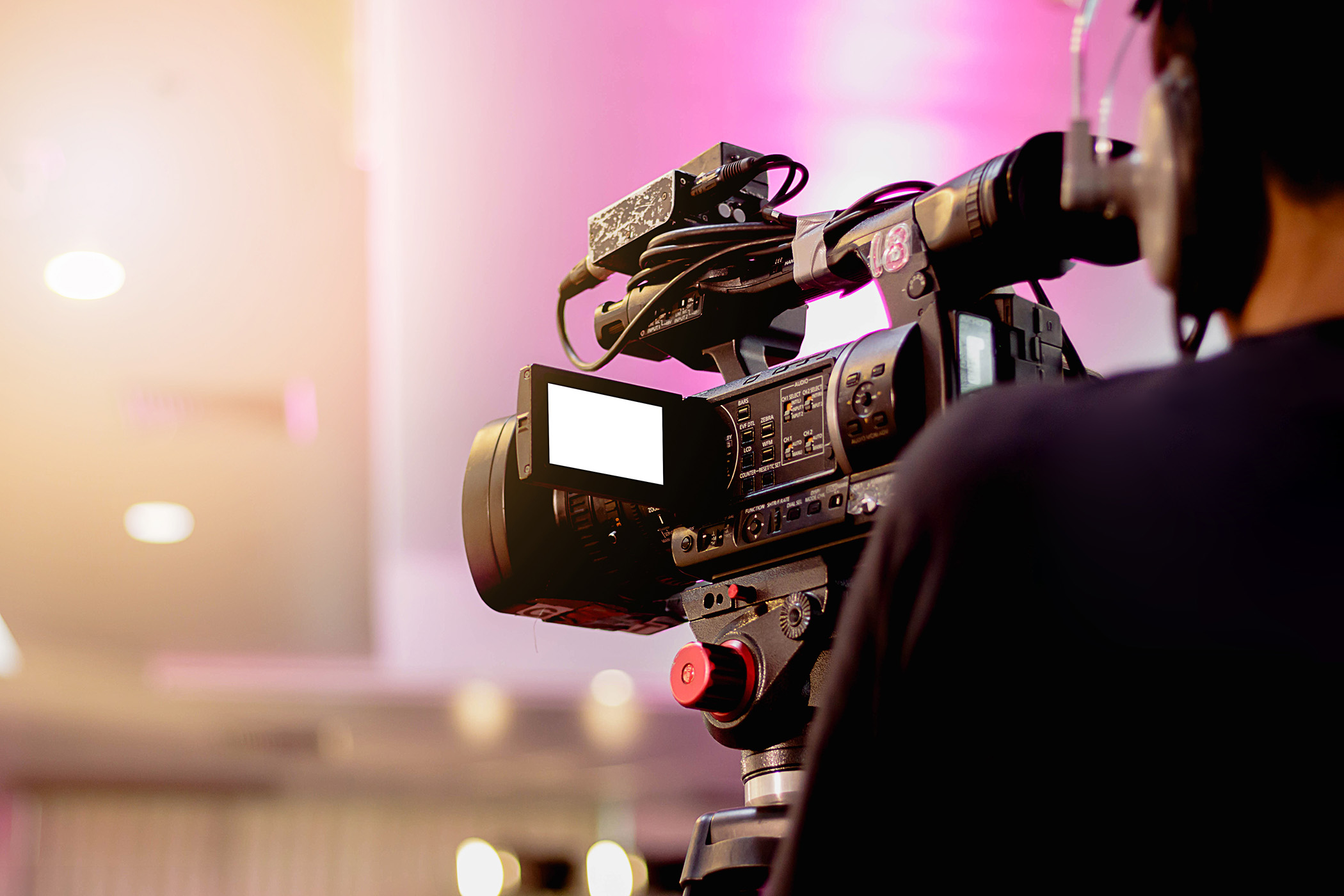 A professional video camera operated by a person in a studio or event setting, with a blurred background and pink lighting.