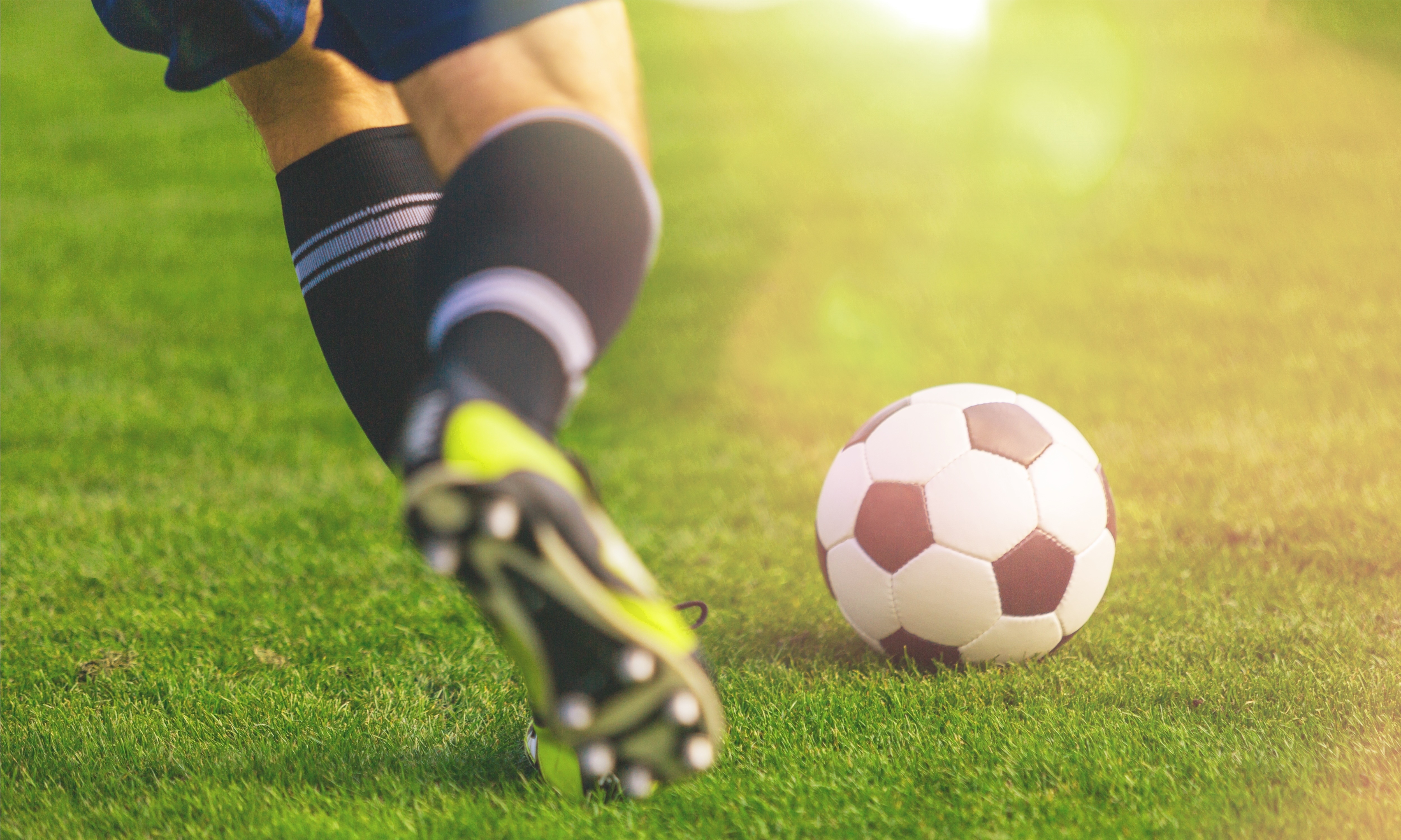A close-up of a soccer player in motion, wearing black socks and yellow cleats, about to kick a black and white soccer ball on a bright green grass field, with sunlight illuminating the scene.