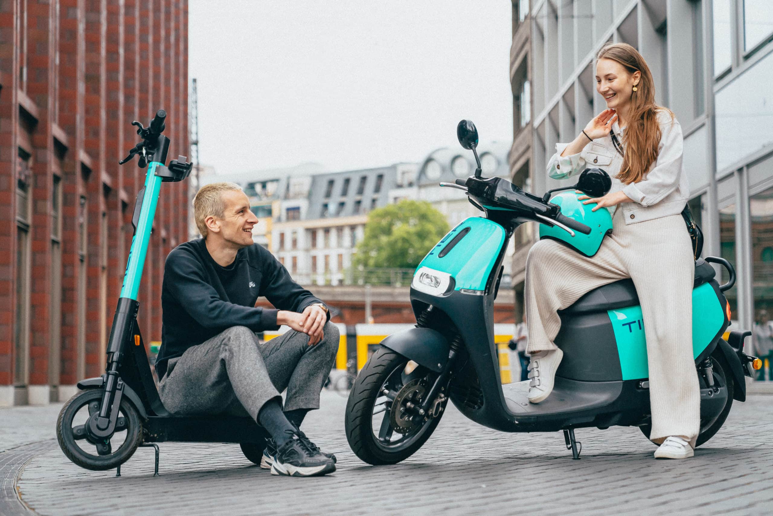A man sits on an electric scooter while talking to a woman seated on an electric moped in a city environment. Both personal mobility vehicles are turquoise and black, and the scene highlights modern urban transportation and friendly interaction.