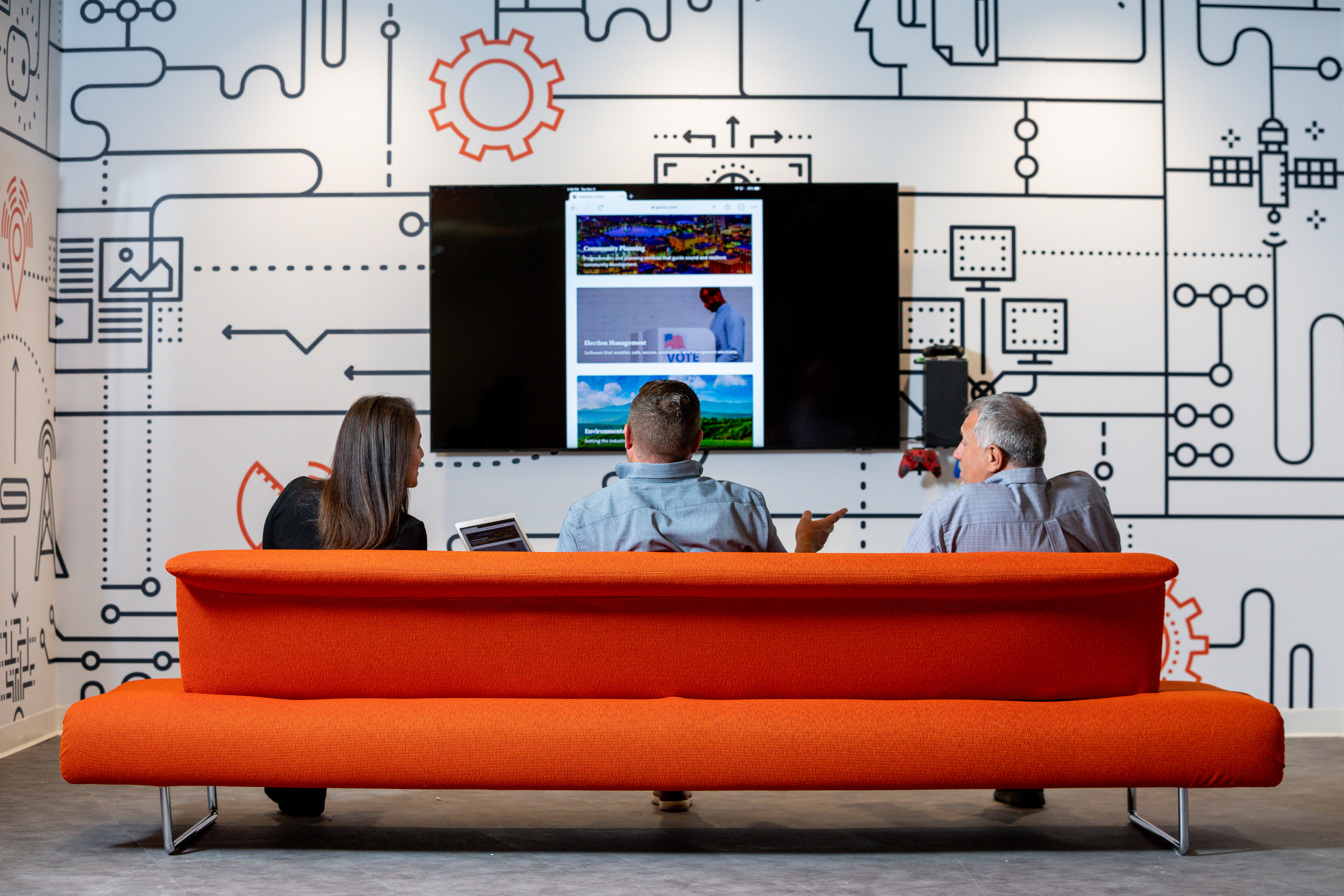 Three people sitting on an orange couch facing a large screen in a modern office space, discussing content displayed on the screen. The room features a wall decorated with technology and network-themed graphics.