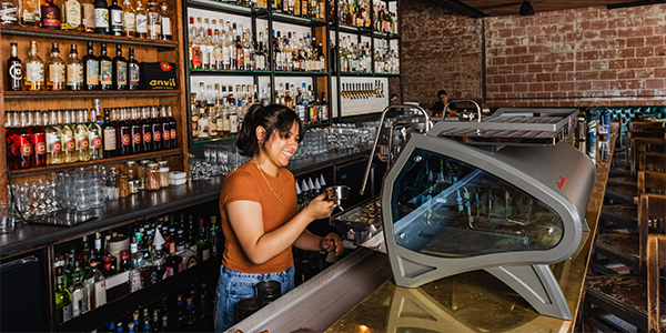 A barista preparing coffee behind the counter of a cocktail bar, surrounded by bottles and glassware.