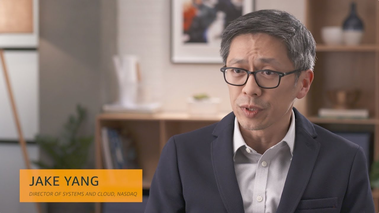 A professional man wearing glasses and a suit is speaking in an office setting. Text overlay reads 'Jake Yang, Director of Systems and Cloud, NASDAQ.' Office shelves and plants are visible in the background.