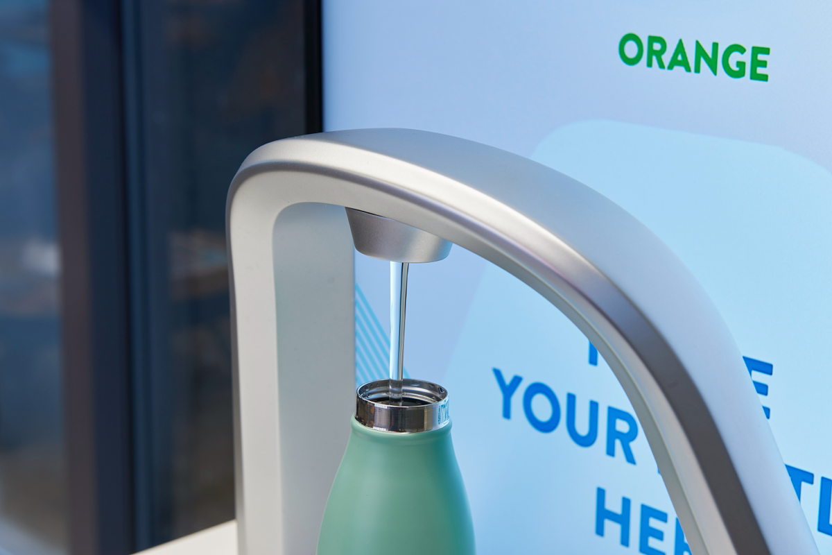 A close-up image of a reusable water bottle being filled with water from a modern bottle refill station. The background includes the word 'ORANGE' and partial signage suggesting eco-friendly messaging.