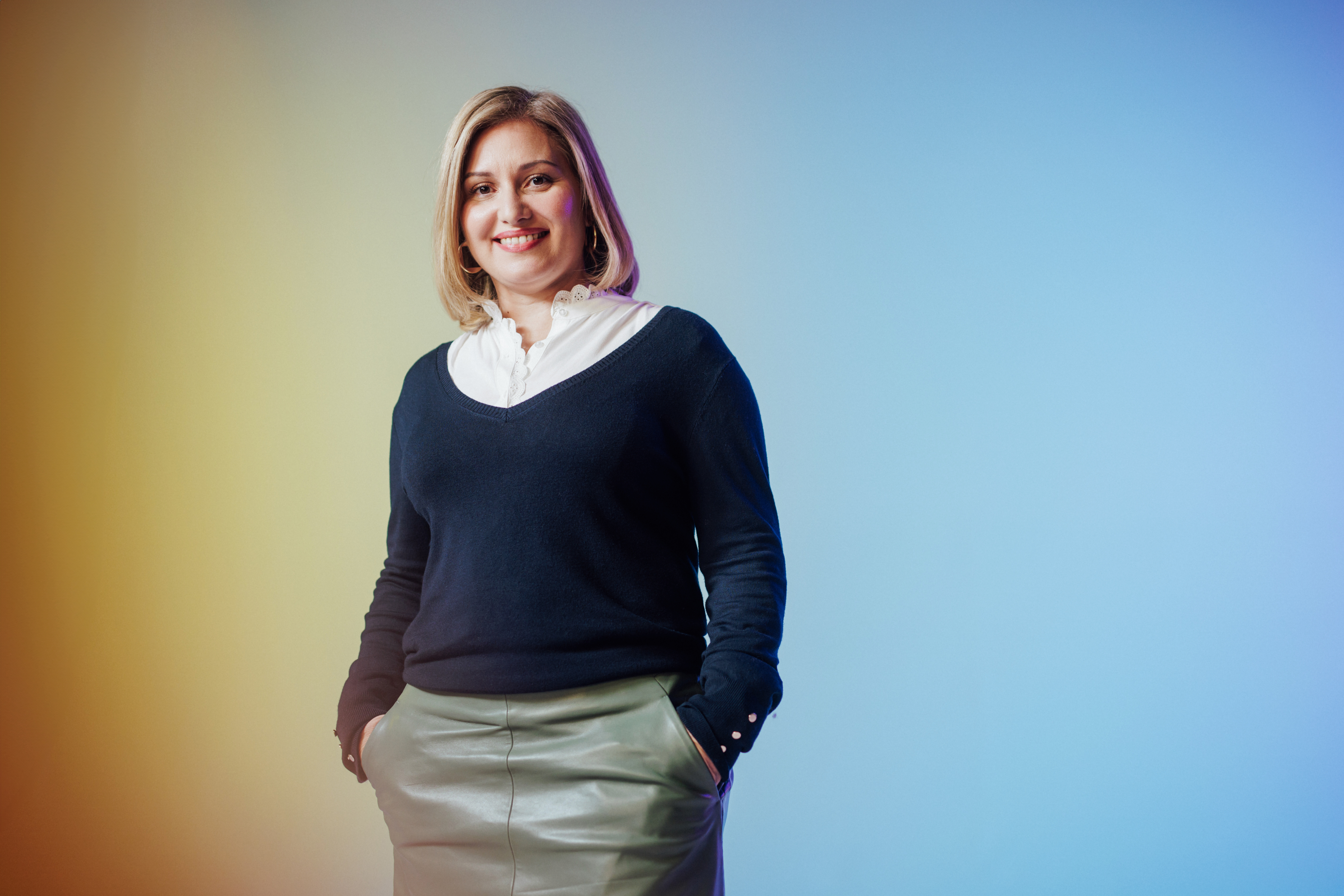 A woman is smiling with her hands in her pockets, standing against a blue and yellow gradient background.